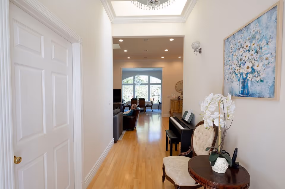 Bright interior hallway with hardwood floors leading into a living and dining area, featuring a piano, vintage chairs, a small round table with flowers, and floral wall art.