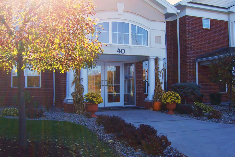 Entrance of a brick building with white columns and double glass doors under an archway with the number 40 above. There are potted yellow flowers and autumn decorations including cornstalks and a pumpkin on either side of the entrance. A tree with yellow and green leaves is visible on the left side with sunlight shining through.