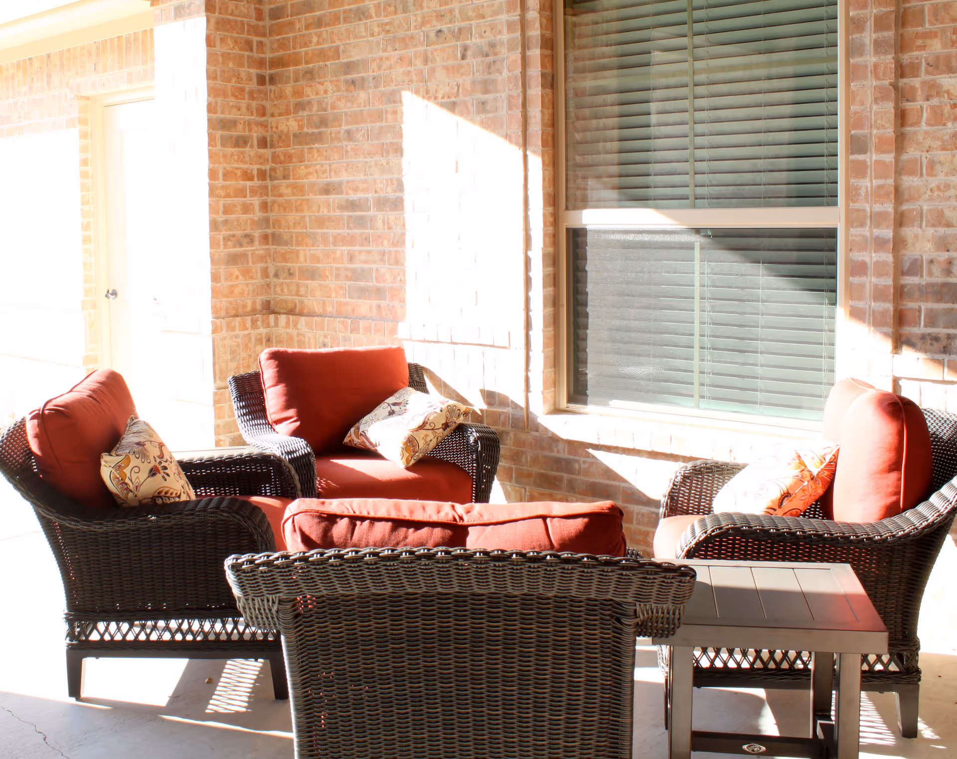 Outdoor seating area with four wicker chairs featuring red cushions and patterned throw pillows arranged around a small wooden table, set against a brick wall with a window and a door nearby.