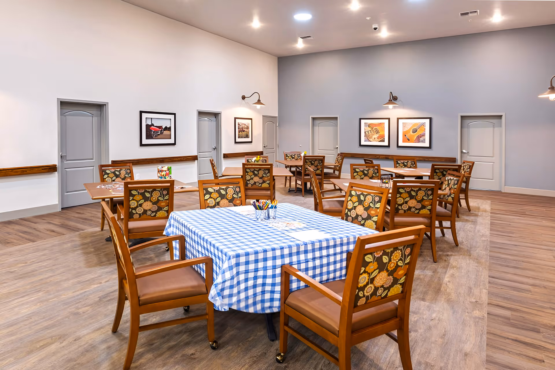 A spacious dining room with multiple wooden tables and chairs. One table in the foreground is covered with a blue and white checkered tablecloth and has containers with colored pencils and papers on it. The chairs have floral-patterned upholstery. The walls are painted in light gray and white, decorated with framed pictures and wall-mounted lamps. The floor is wooden, and there are several closed doors along the walls.