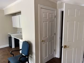 Interior view of a room in a senior living facility showing a corner with two closed white doors, a small built-in desk area with white cabinets above, and a blue office chair on wheels. The walls are painted light beige and the floor is dark wood.