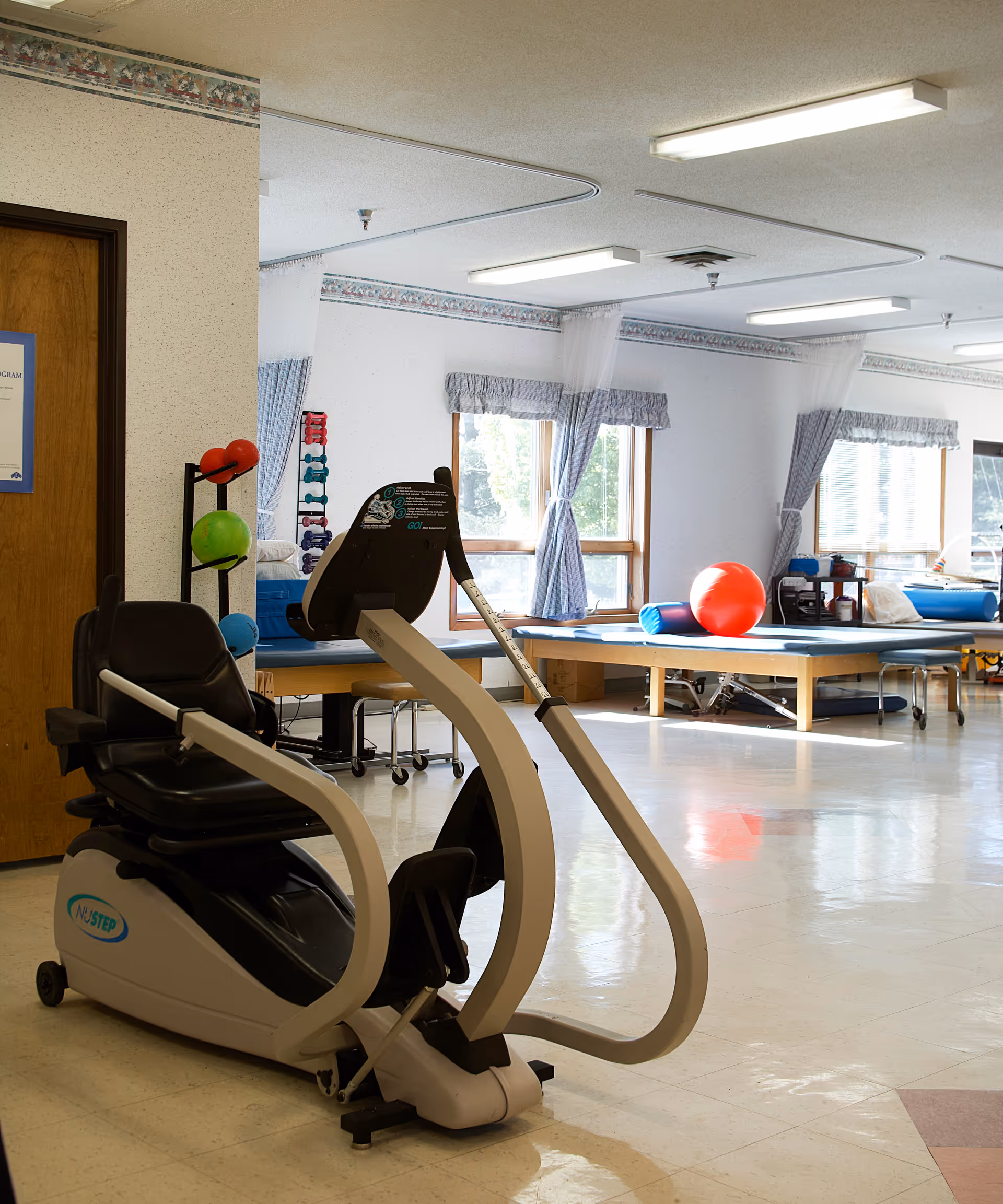 A bright physical therapy or exercise room with a recumbent exercise bike in the foreground, therapy tables with exercise balls and cushions in the background, and windows with blue curtains letting in natural light.