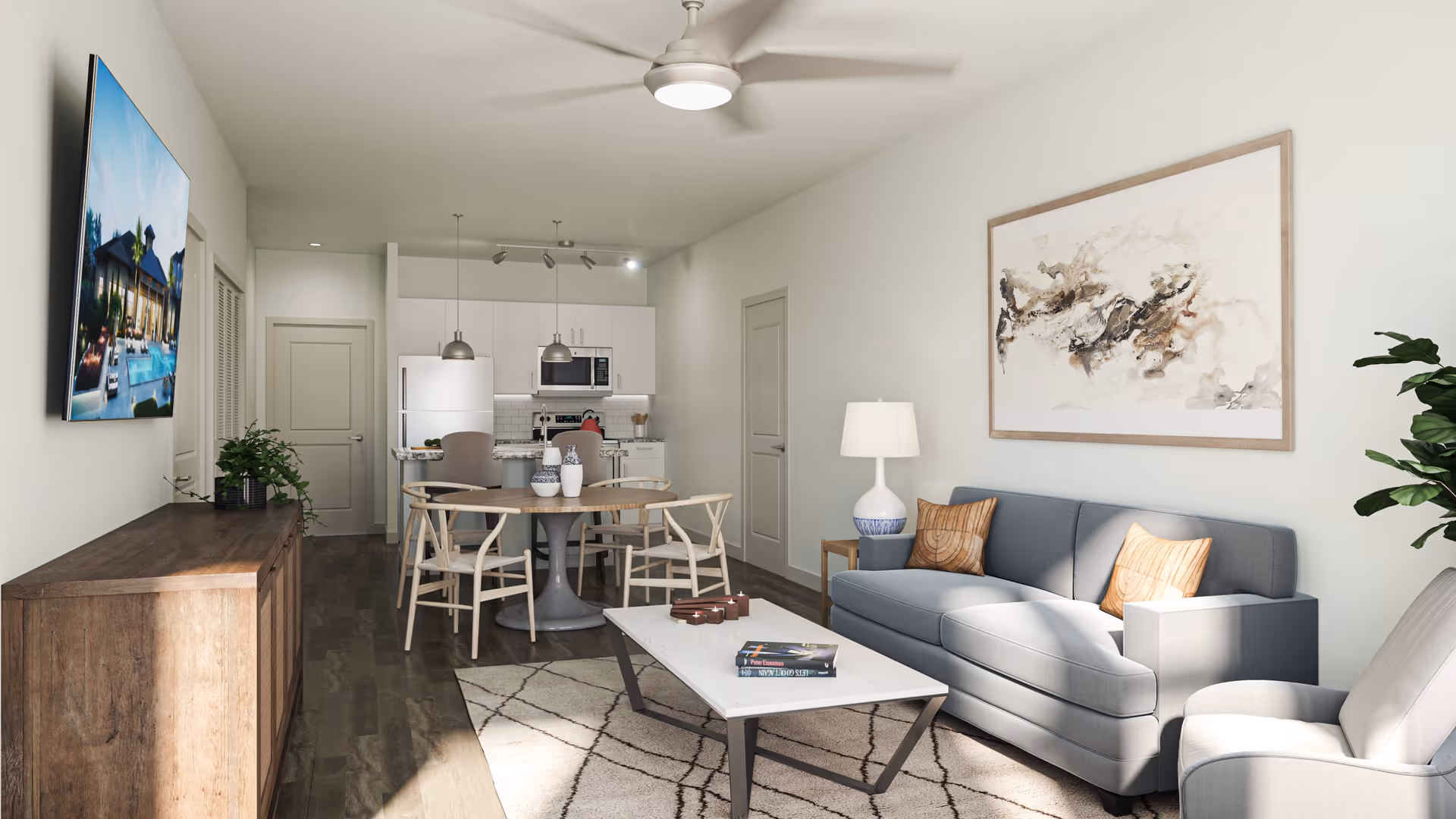 A modern living room and kitchen area with a gray sofa, two wooden chairs around a round dining table, a white armchair, a coffee table with books and candles, a wooden sideboard with a plant, a wall-mounted TV, and a ceiling fan. The kitchen features white cabinets, a refrigerator, a microwave, and pendant lights.