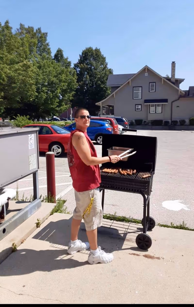 A person wearing sunglasses, a red sleeveless shirt, beige shorts, and white sneakers is grilling sausages on a black barbecue grill outdoors in a parking lot. There are several parked cars and a house with trees in the background.