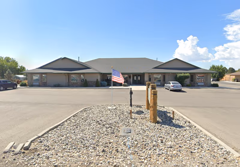Front exterior view of a single-story assisted living facility building with a peaked roof, beige siding, and stone accents. There is a paved parking lot in front with a few parked cars, a small landscaped area with rocks, wooden posts, and an American flag on a flagpole. The sky is clear with some clouds.
