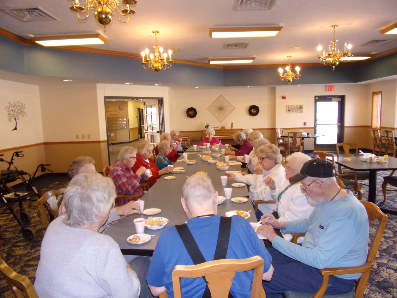 A group of elderly residents seated around a long table eating and socializing in a senior living dining room.