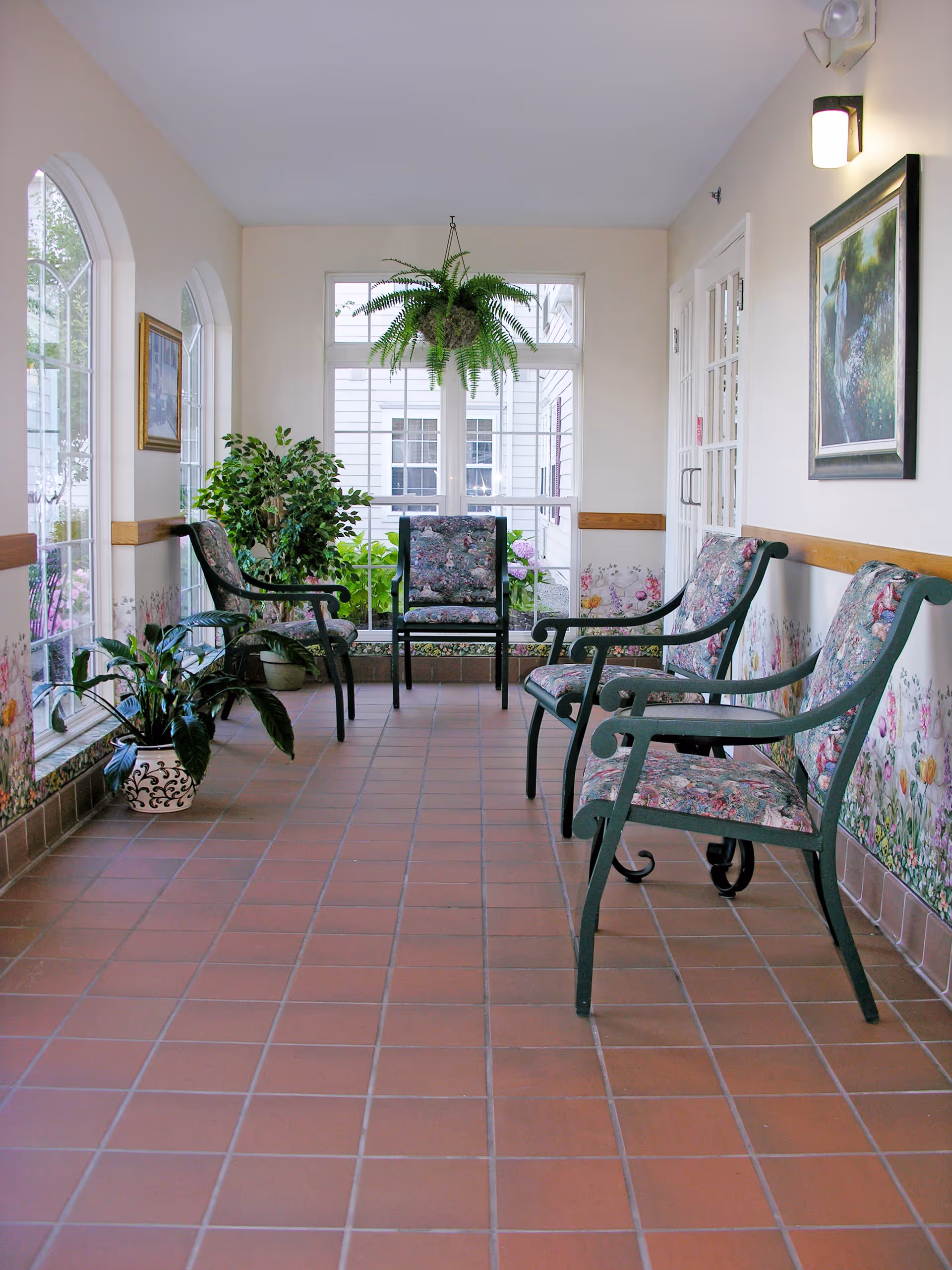 A bright indoor seating area with floral-patterned chairs arranged along the walls and a hanging fern plant in front of large windows. The floor is tiled with reddish-brown tiles, and there are several potted plants placed near the windows. The walls are decorated with floral wallpaper and framed artwork.