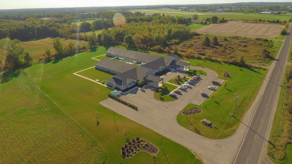 Aerial view of a single-story senior living facility named Quality Care of Howell, surrounded by green lawns, trees, and open fields. The building has a large parking lot with several cars parked, and a road runs alongside the property.