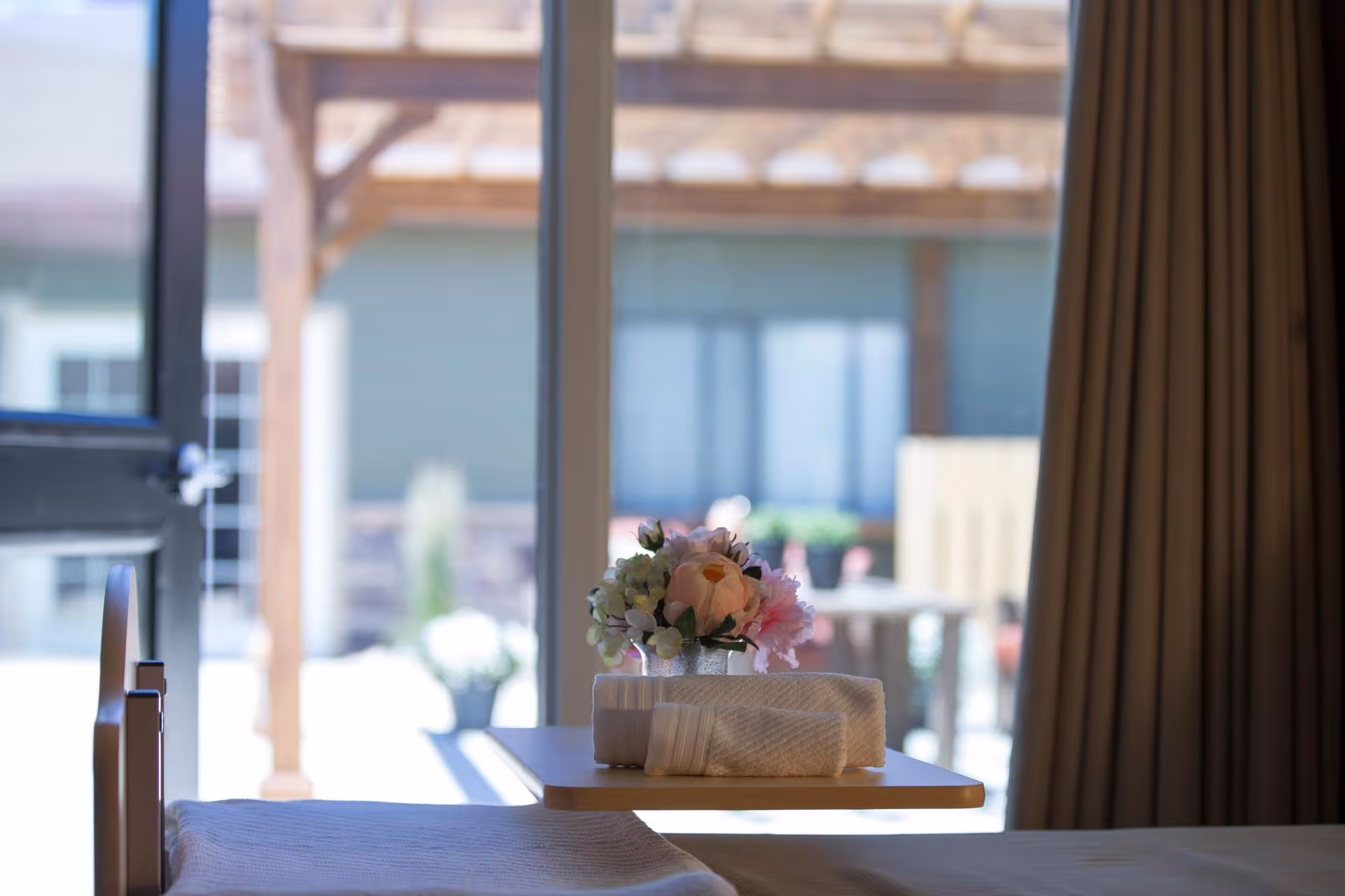 A close-up view of a bedside table with a small bouquet of flowers and neatly folded towels, with a sliding glass door and outdoor patio visible in the background.