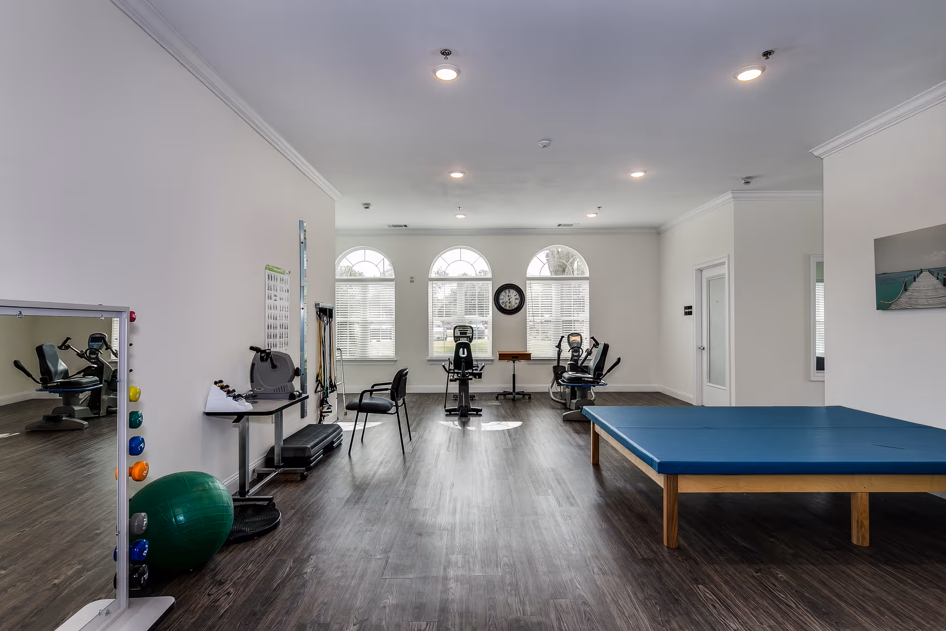 A bright exercise room with wooden flooring and white walls featuring three large arched windows with blinds. The room contains exercise equipment including stationary bikes, a green exercise ball, resistance bands, and a blue padded table. There is a wall clock between the windows and a framed picture on the right wall.
