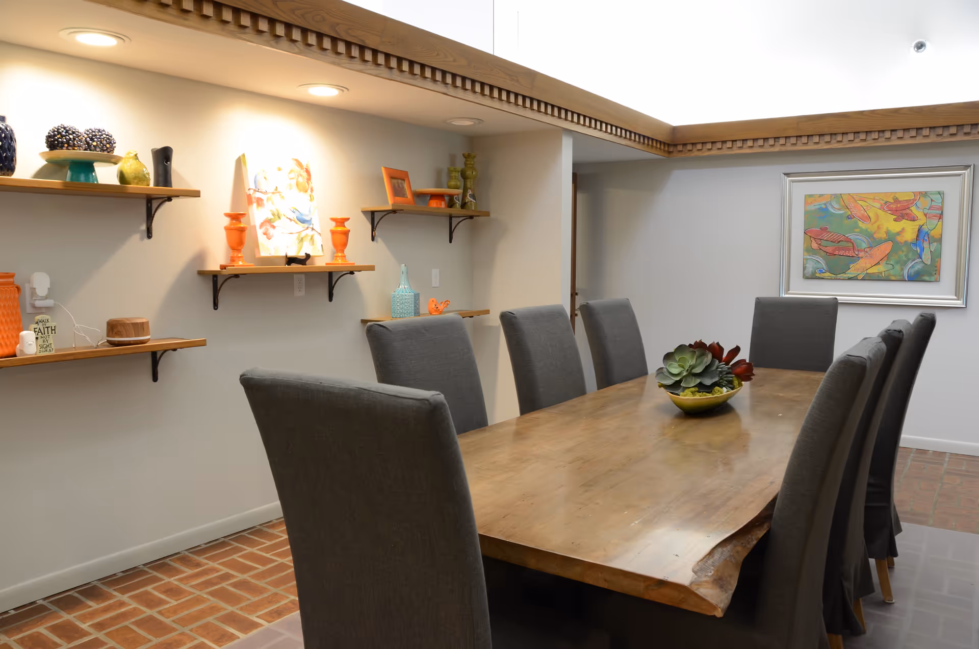 A dining room with a long wooden table surrounded by eight gray upholstered chairs. The table has a decorative bowl with succulents in the center. The walls are light-colored with wooden shelves holding various decorative items and a colorful framed painting. The floor is tiled with a brick pattern.
