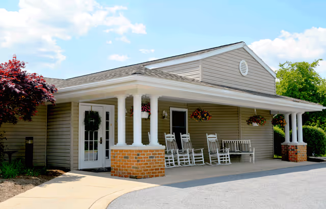 Exterior view of a single-story building with beige siding and a covered porch supported by white columns with brick bases. The porch has several white rocking chairs and hanging flower baskets. There is a paved driveway and a small landscaped area with a tree and bushes under a partly cloudy sky.