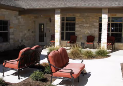 Outdoor patio area with cushioned metal chairs and benches arranged around a circular garden bed, adjacent to a stone building with windows and columns.