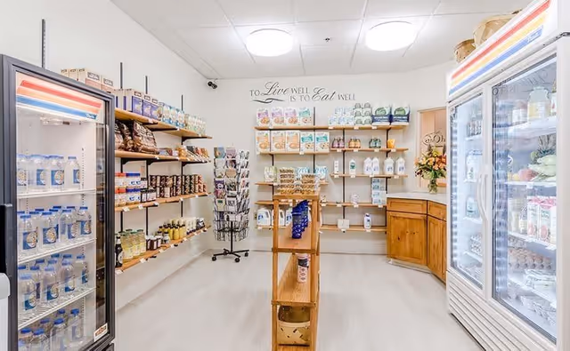 Interior view of a small grocery or convenience store area with shelves stocked with various food and household items. Two glass-door refrigerators are visible on the left and right sides, containing beverages and other refrigerated products. Wooden shelves in the center and along the walls hold packaged goods, snacks, and other items. A sign on the back wall reads 'To Live Well is to Eat Well.'