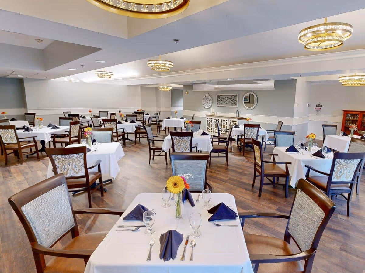 A spacious dining room with multiple tables covered in white tablecloths, each set with navy blue napkins, glassware, and silverware. The chairs have wooden frames with patterned upholstery. The room features wooden flooring, soft lighting from modern circular chandeliers, and decorative wall elements including a clock and a mirror.