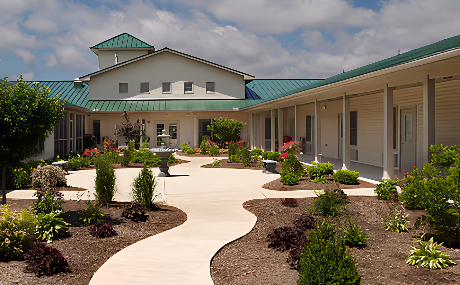 Landscaped courtyard with a winding concrete path leading to a single-story senior living building with green metal roofs.