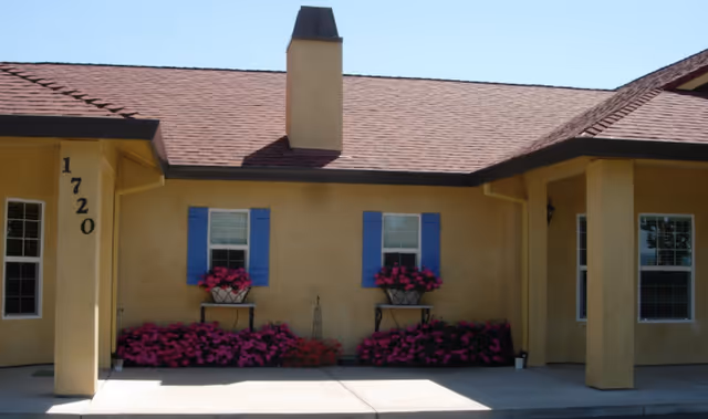 Front facade of a single-story beige stucco building with a red roof, two blue-shuttered windows and pink flowers beneath, and the number 1720 on a column.