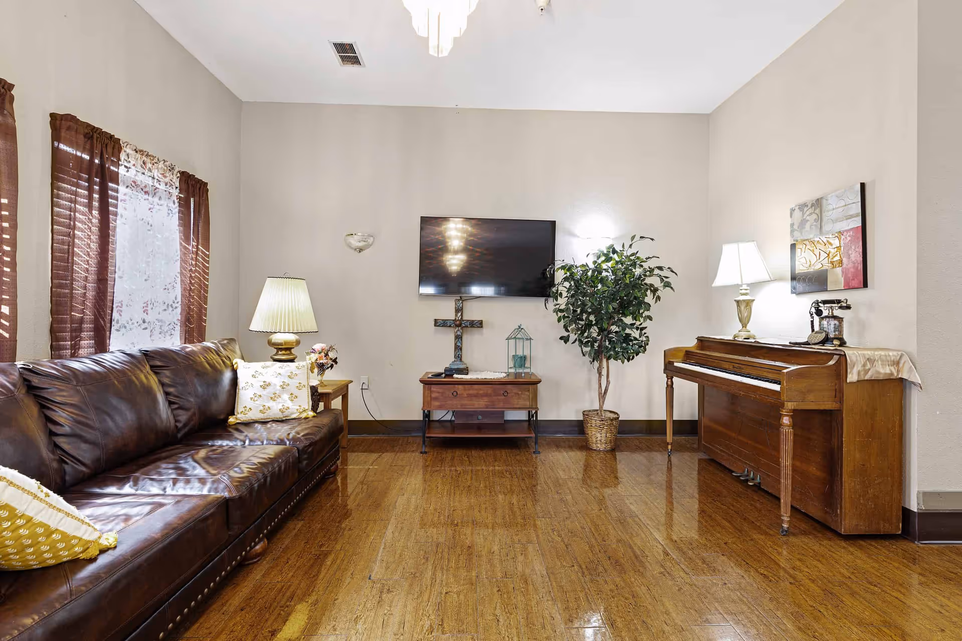 A cozy living room with a dark brown leather sofa on the left, a wooden piano on the right, and a small wooden table with a cross and decorative items in the center beneath a wall-mounted flat-screen TV. There are two table lamps on either side of the room, a potted plant next to the piano, and windows with brown curtains on the left wall.