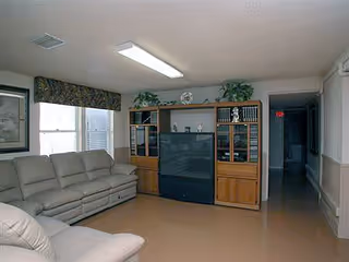 A living room with beige leather sofas arranged around a large wooden entertainment center holding a large television. The room has two windows with floral valances, a framed picture on the wall, and a hallway leading to other rooms.
