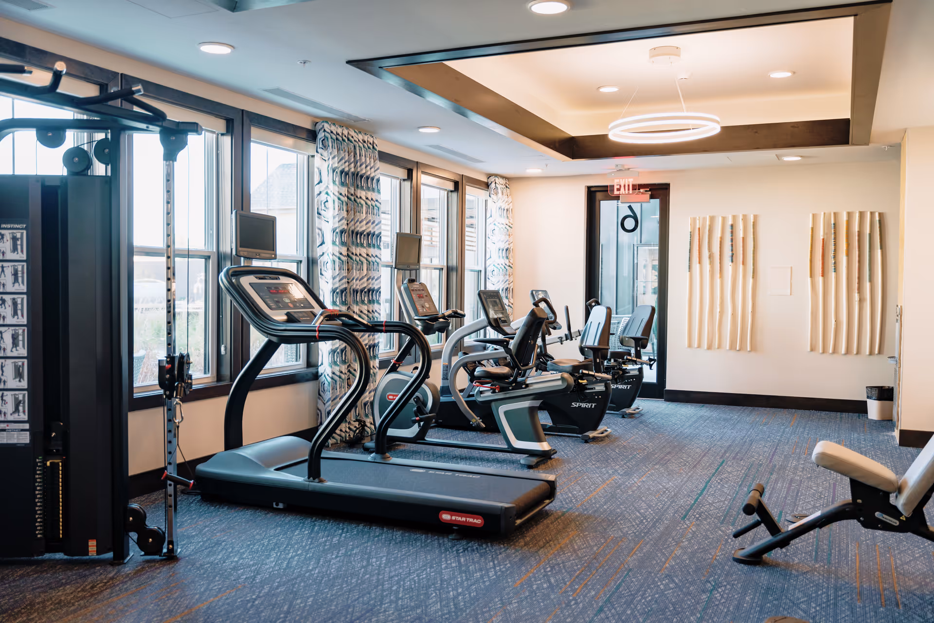 A well-lit fitness room with exercise equipment including treadmills, stationary bikes, and a weight machine. Large windows with patterned curtains allow natural light to fill the space. The room has a modern ceiling light fixture and decorative wall art.
