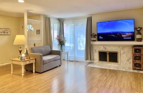 A bright living room with light wood flooring, a gray loveseat, a wooden side table with a lamp and decorative items, a white marble fireplace with a flat-screen TV mounted above it, and large windows with curtains letting in natural light.