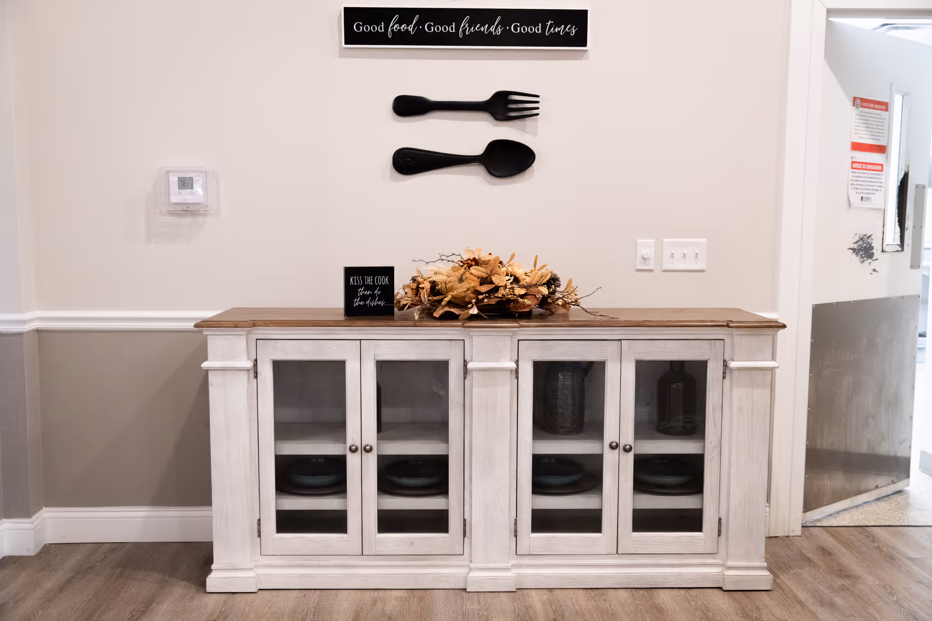 A white wooden cabinet with glass doors displaying dishes inside, topped with a decorative arrangement of dried flowers and a small sign that reads 'Kiss the cook, clean up the dishes.' Above the cabinet on the wall are large black decorative fork and spoon and a sign that says 'Good food · Good friends · Good times.' The room has light-colored walls with a thermostat on the left and light switches on the right, with a partially open door leading to another area.