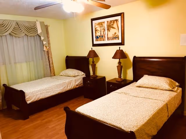 A bedroom with two single beds featuring dark wooden frames and beige patterned bedding. Between the beds is a dark wooden nightstand with two matching table lamps. Above the nightstand hangs a framed picture of palm trees. The room has light-colored walls, a ceiling fan with a light, and a window with decorative curtains.