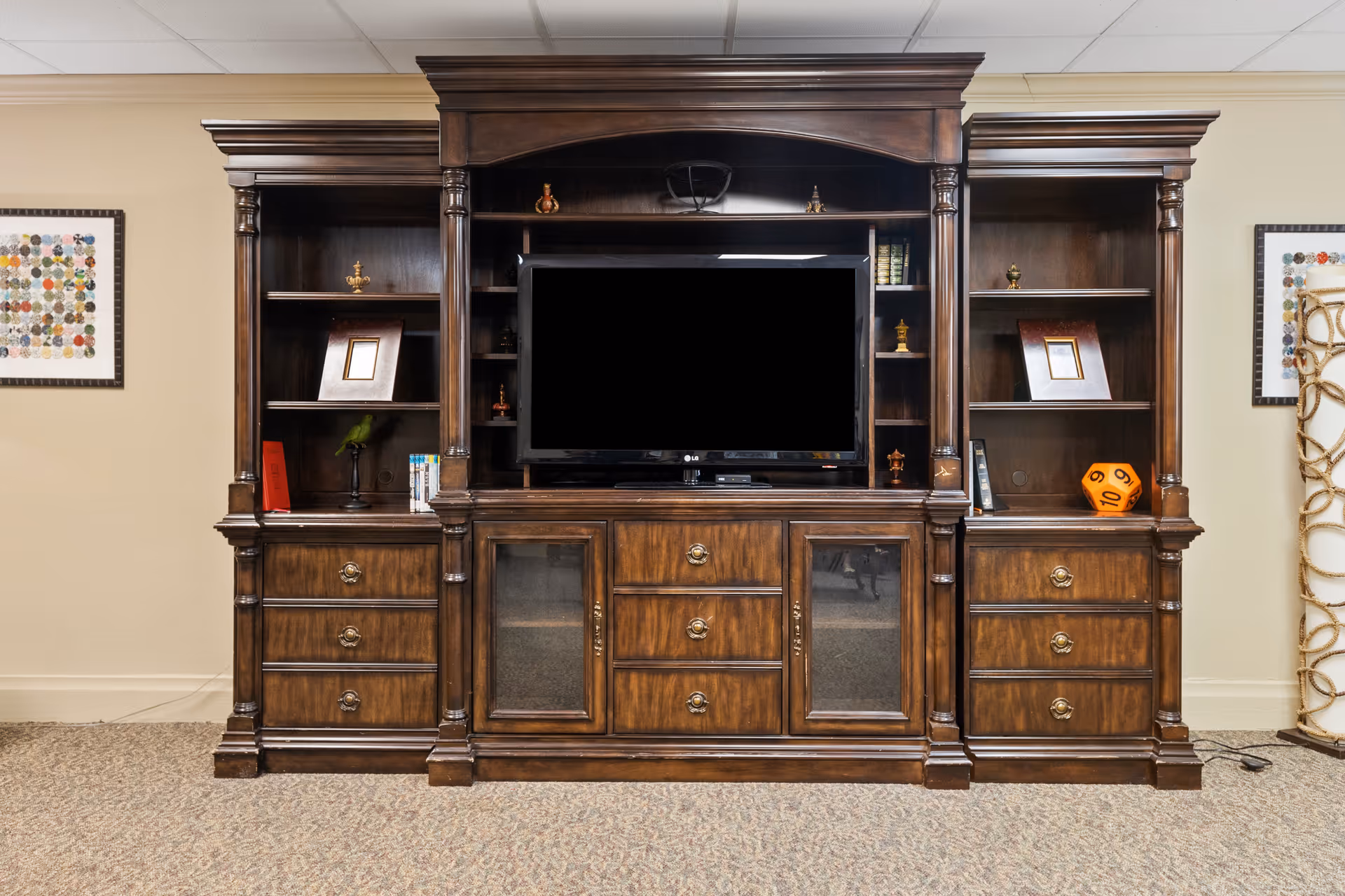 A large wooden entertainment center with multiple shelves and drawers. A flat-screen TV is centered in the middle section. The shelves contain decorative items, books, and picture frames. The unit is placed against a beige wall with framed artwork on either side.