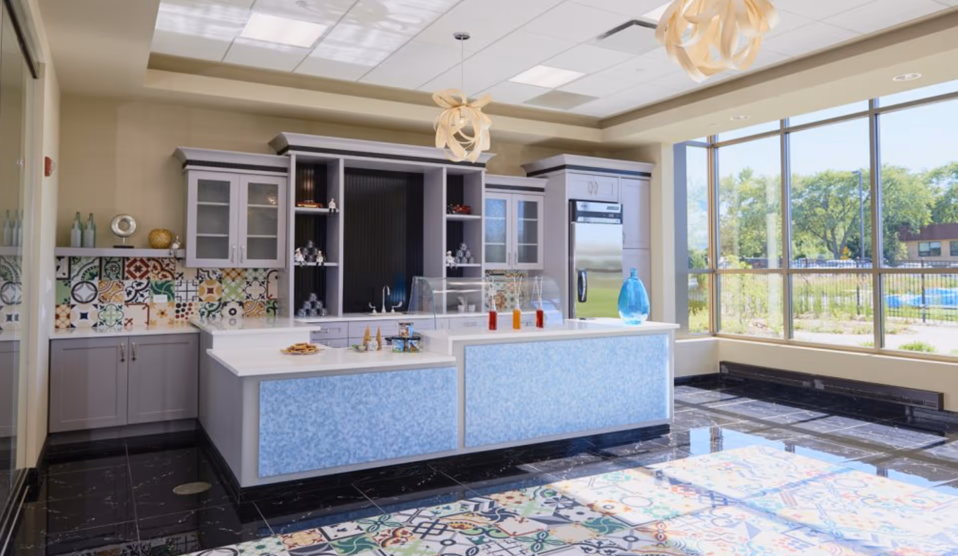 Bright and modern kitchen area with large windows allowing natural light to flood the space. The kitchen features a central island with a blue patterned front, a countertop with various items including bottles and snacks, and cabinets with glass doors. The backsplash has colorful, patterned tiles, and the floor has a mix of black tiles and patterned tiles. Two decorative pendant lights hang from the ceiling.