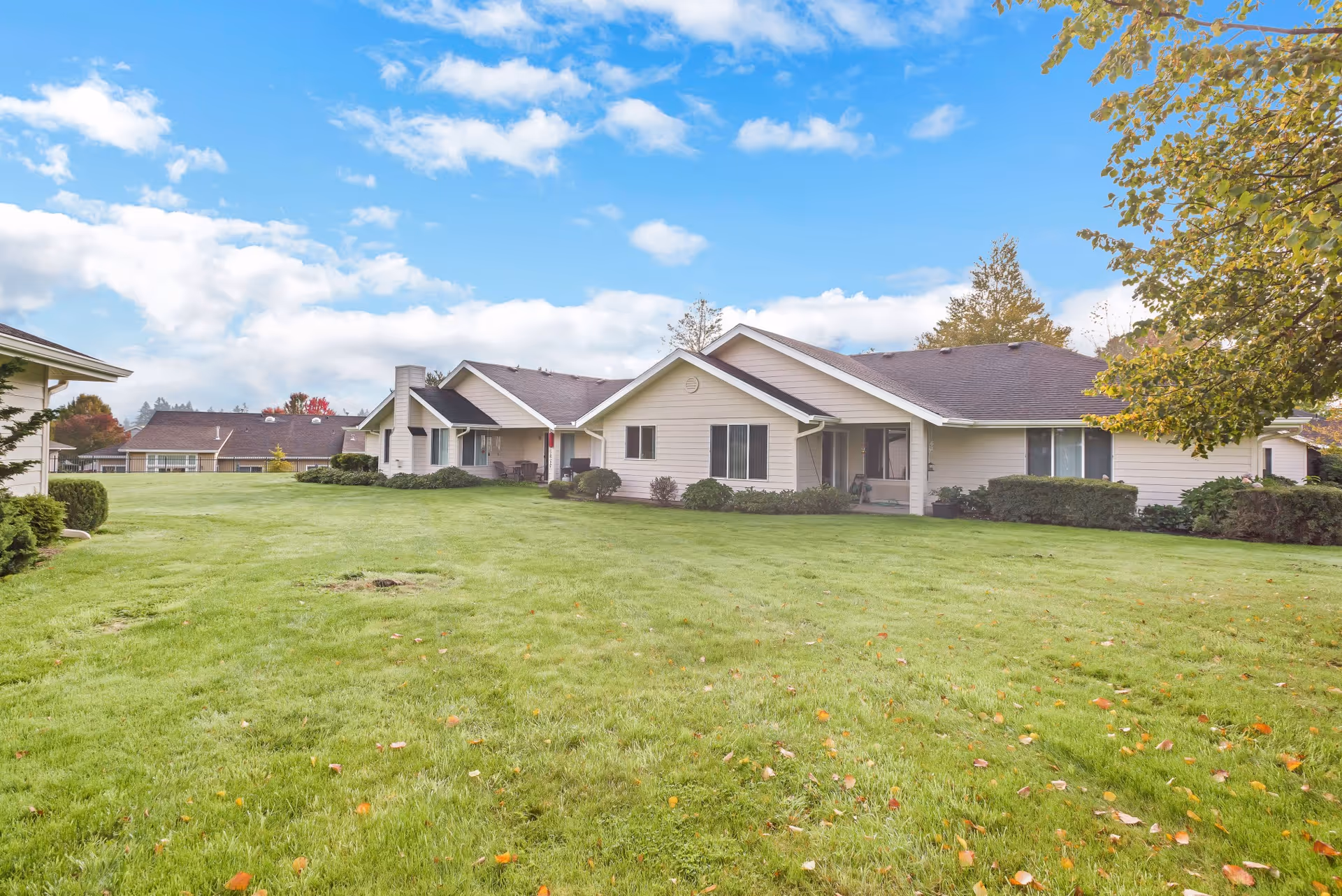 Exterior view of single-story residential buildings with beige siding and dark roofs, surrounded by a well-maintained green lawn and some trees under a partly cloudy blue sky.