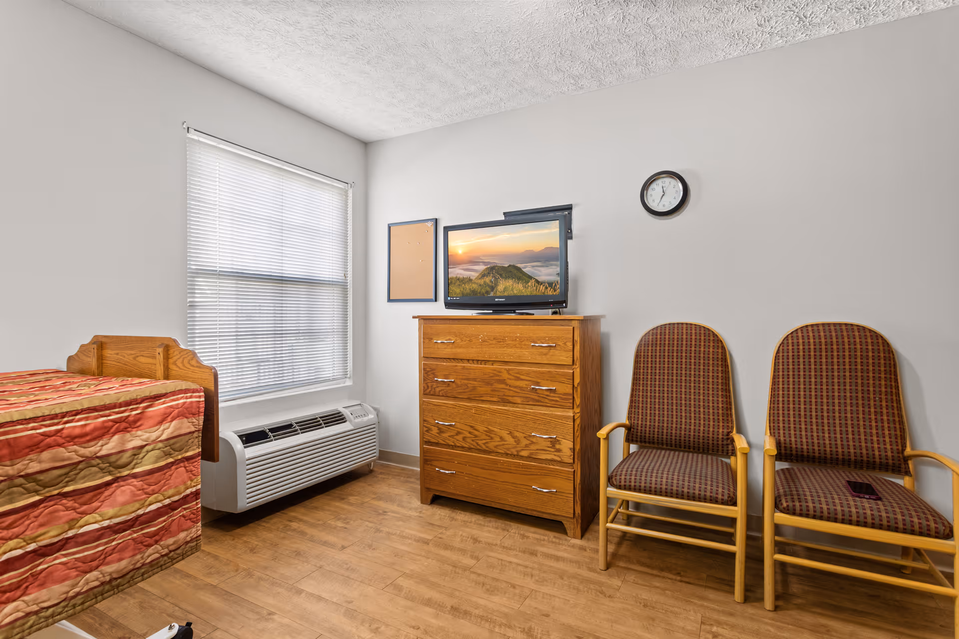 A small room with a single bed covered in a striped red and beige quilt, a wooden dresser with a flat-screen TV on top, two wooden chairs with patterned cushions, a wall clock, a window with blinds, and a heating/cooling unit beneath the window.