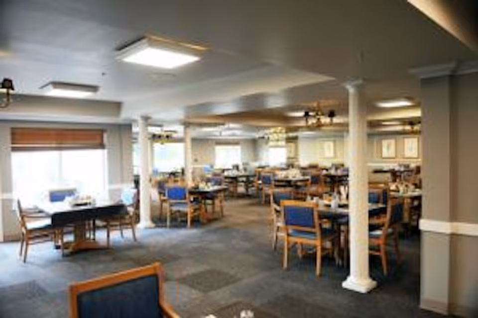 A spacious dining room with multiple wooden tables and chairs with blue cushions. The room features large windows with blinds, white columns, and ceiling lights providing bright illumination. The tables are set with napkins and utensils, creating a welcoming atmosphere.
