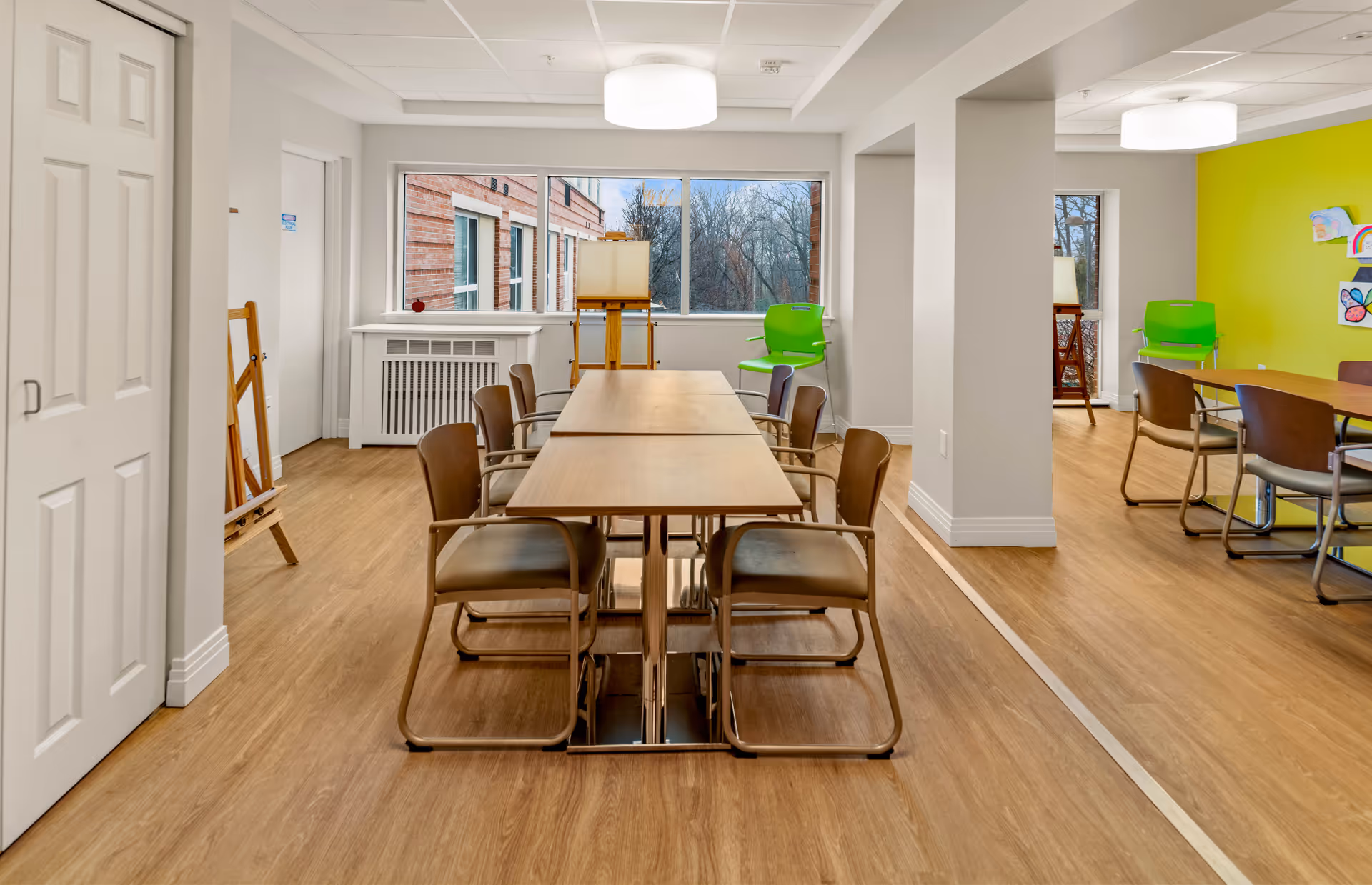 Bright communal activity room with long tables and chairs, wood floors, large windows and a green accent wall.