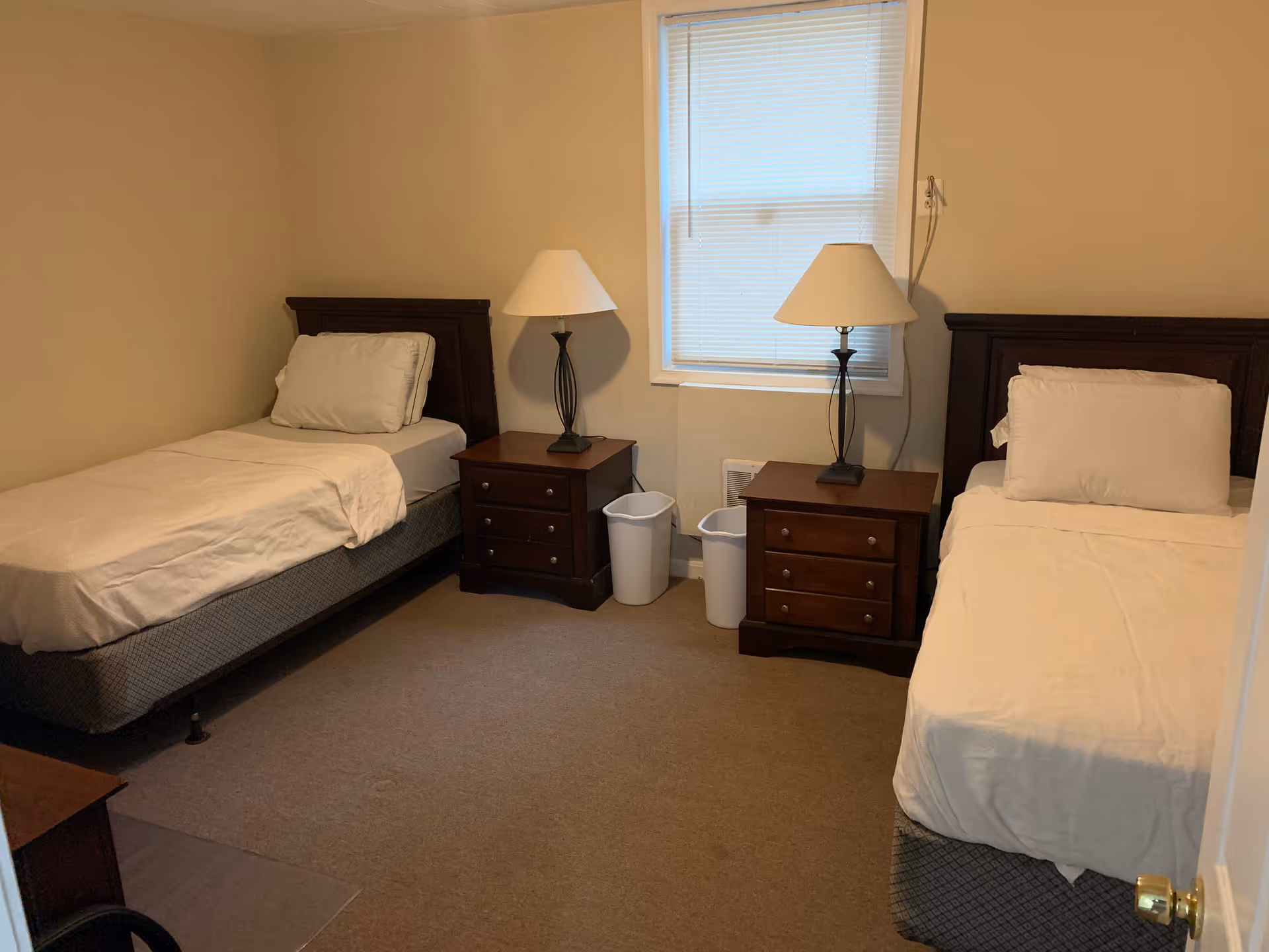 A small bedroom with two single beds, each with white bedding and a pillow. Between the beds are two dark wooden nightstands, each with a lamp. There is a window with closed blinds above the nightstands. Two white trash bins are placed between the nightstands. The walls are beige and the floor is carpeted.