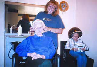 An elderly woman sitting in a salon chair smiling while a hairdresser stands behind her, styling her hair. In the background, another elderly woman is seated under a hair dryer, reading a magazine.