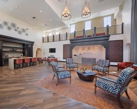 Spacious two-story community lounge with patterned armchairs around a low coffee table, a dining/bar area to the left, and a mezzanine balcony under high ceilings.