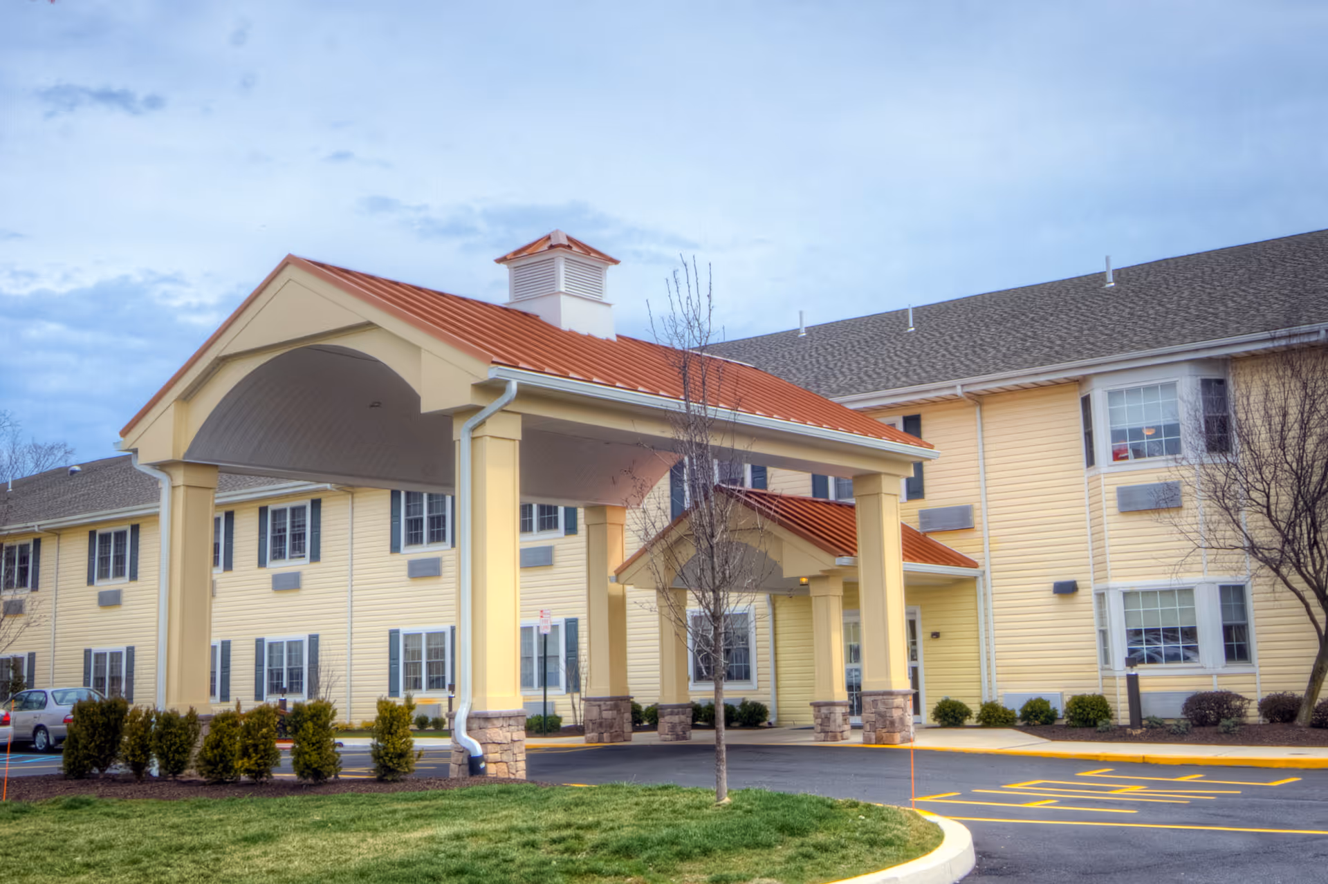 Exterior view of a senior living facility building with yellow siding and a red roof. The entrance has a covered drop-off area supported by columns. There are small bushes and a tree in front of the building, and a parking lot with marked spaces is visible.