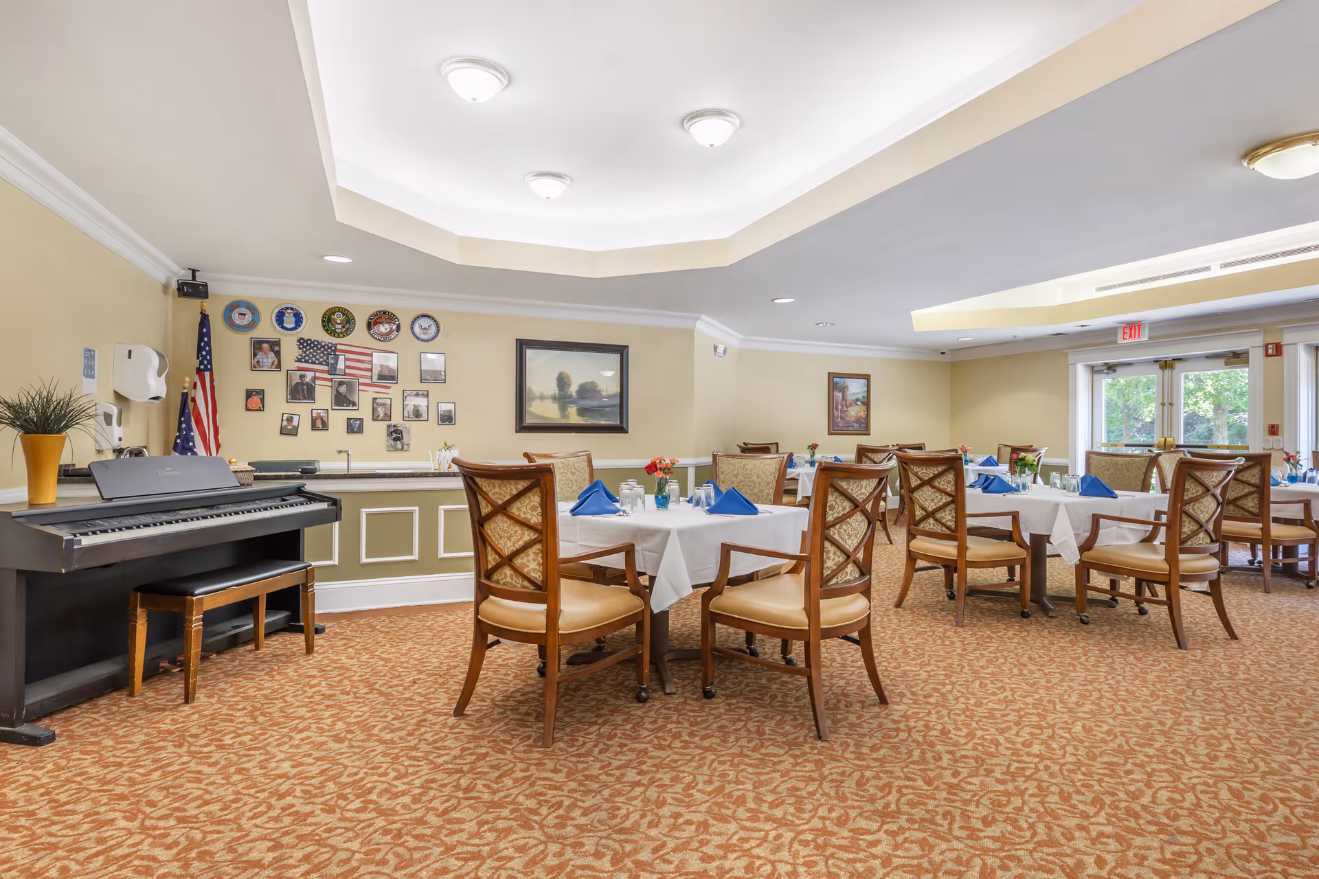 A dining room in a senior living facility with several tables covered with white tablecloths, each set with blue napkins, glasses, and small flower arrangements. The room has beige walls adorned with framed pictures and military emblems. There is a piano with a bench on the left side and an American flag next to it. Large windows and glass doors allow natural light to enter the room.