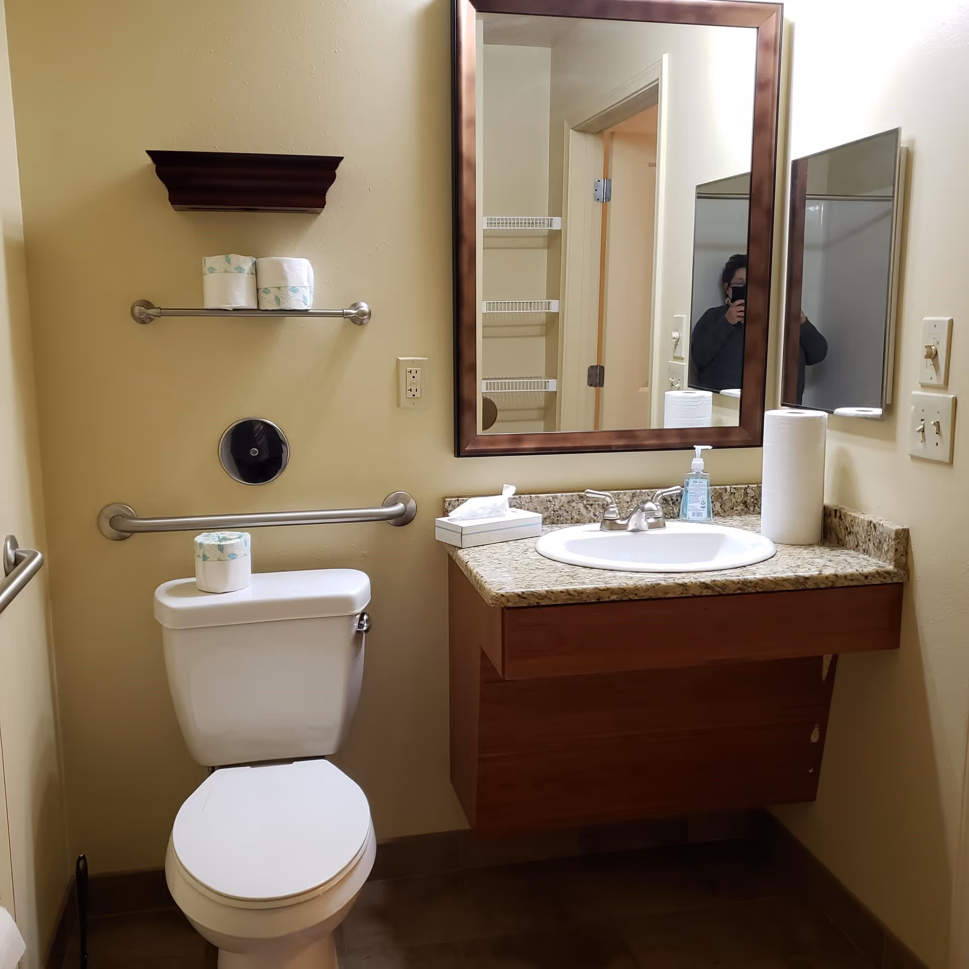A bathroom with a white toilet, a granite countertop with a sink, a large rectangular mirror with a brown frame, a paper towel roll, a soap dispenser, and a tissue box. There are two metal grab bars on the wall, a small shelf with two rolls of toilet paper, and a person partially visible in the mirror taking the photo.