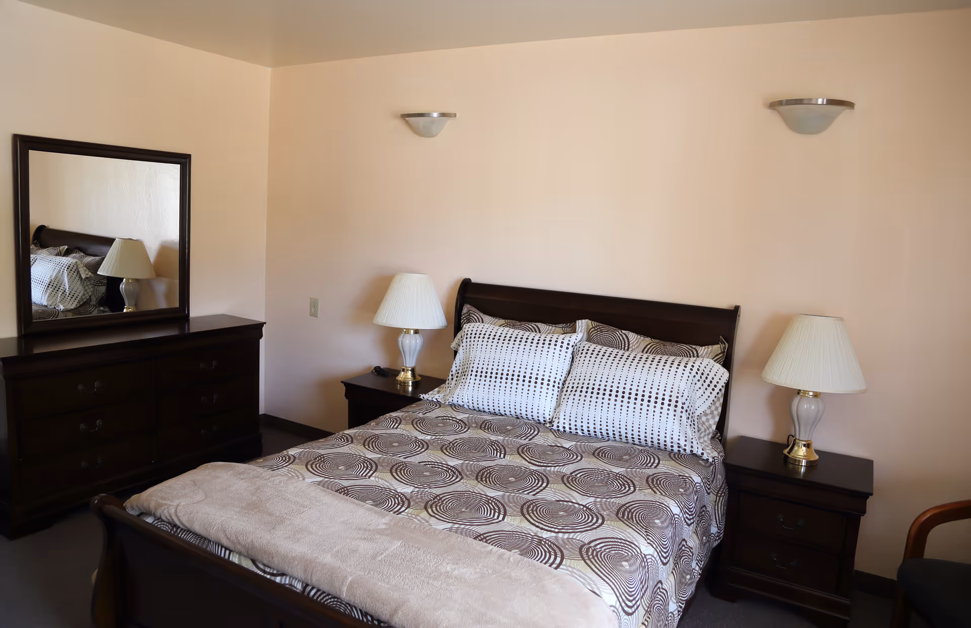 A neatly made bed with patterned bedding and two pillows in a bedroom. The bed has a dark wooden frame and is flanked by two matching nightstands, each with a white lamp. To the left, there is a dark wooden dresser with a large mirror above it. The walls are painted light beige, and there are two wall-mounted light fixtures above the bed.