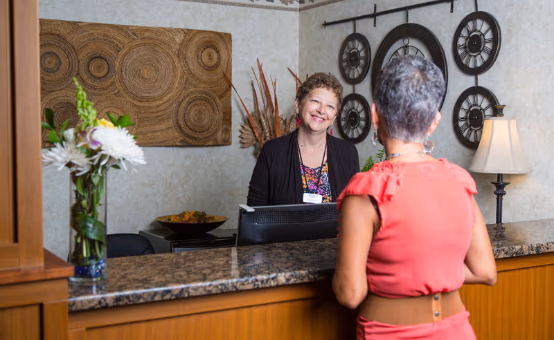 A smiling receptionist standing behind a granite countertop desk talking to a woman in a coral dress at the front desk of a facility. The background features decorative wall art, a lamp, and a vase with flowers on the counter.