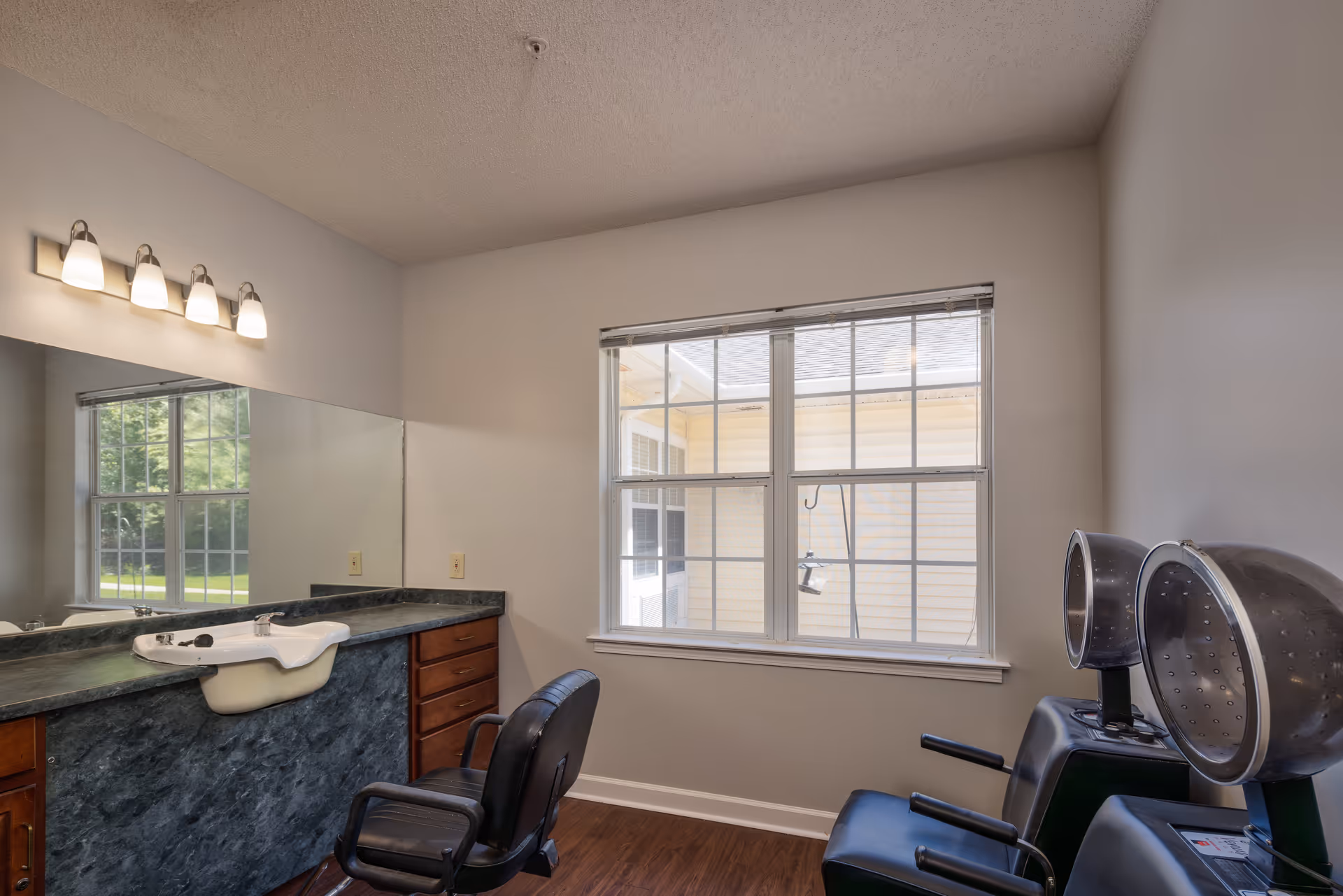Interior view of a hair salon area in an assisted living facility featuring a large mirror above a countertop with a built-in sink, a black salon chair, and two hair dryer chairs near a window with blinds.