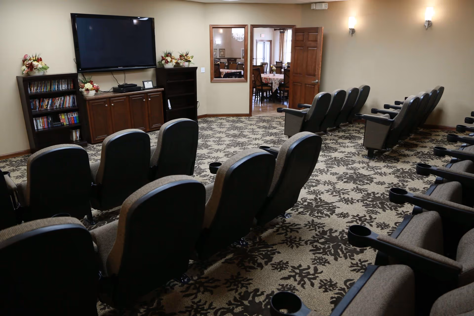 A small theater room with two rows of cushioned chairs facing a large flat-screen TV mounted on the wall. There are bookshelves and cabinets beneath the TV with decorative flower arrangements on top. The room has patterned carpet and beige walls with wall-mounted lights. An open door leads to a dining area with tables and chairs.