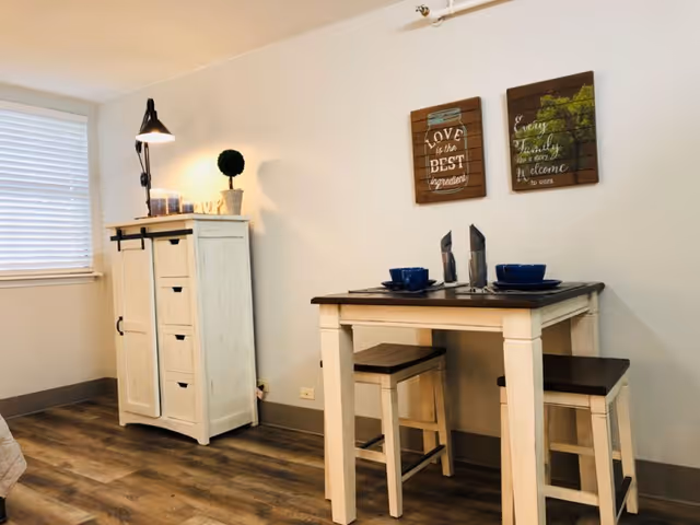 A small dining area in a room with a wooden table and two matching stools. The table is set with blue bowls and napkins. On the wall above the table are two decorative wooden plaques with inspirational quotes. To the left, there is a white cabinet with drawers and a lamp on top, next to a window with closed blinds. The floor has wood-style flooring.