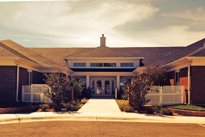 Front exterior view of a single-story brick building with a pitched roof, a central entrance with double glass doors, and a concrete walkway leading up to it. There are small bushes and white picket fences on either side of the entrance.