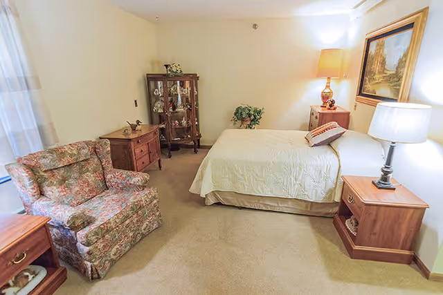 A cozy bedroom with a bed covered in a light-colored quilt, two wooden nightstands with lamps on either side, a floral upholstered armchair, a wooden dresser, and a glass display cabinet in the corner. There is a framed painting on the wall and a potted plant near the bed.