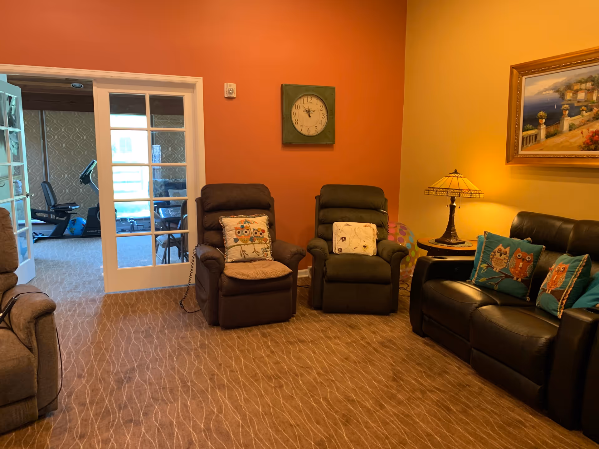 Cozy living room with recliner chairs and a leather sofa around a side table and lamp, with glass doors leading to an exercise room.