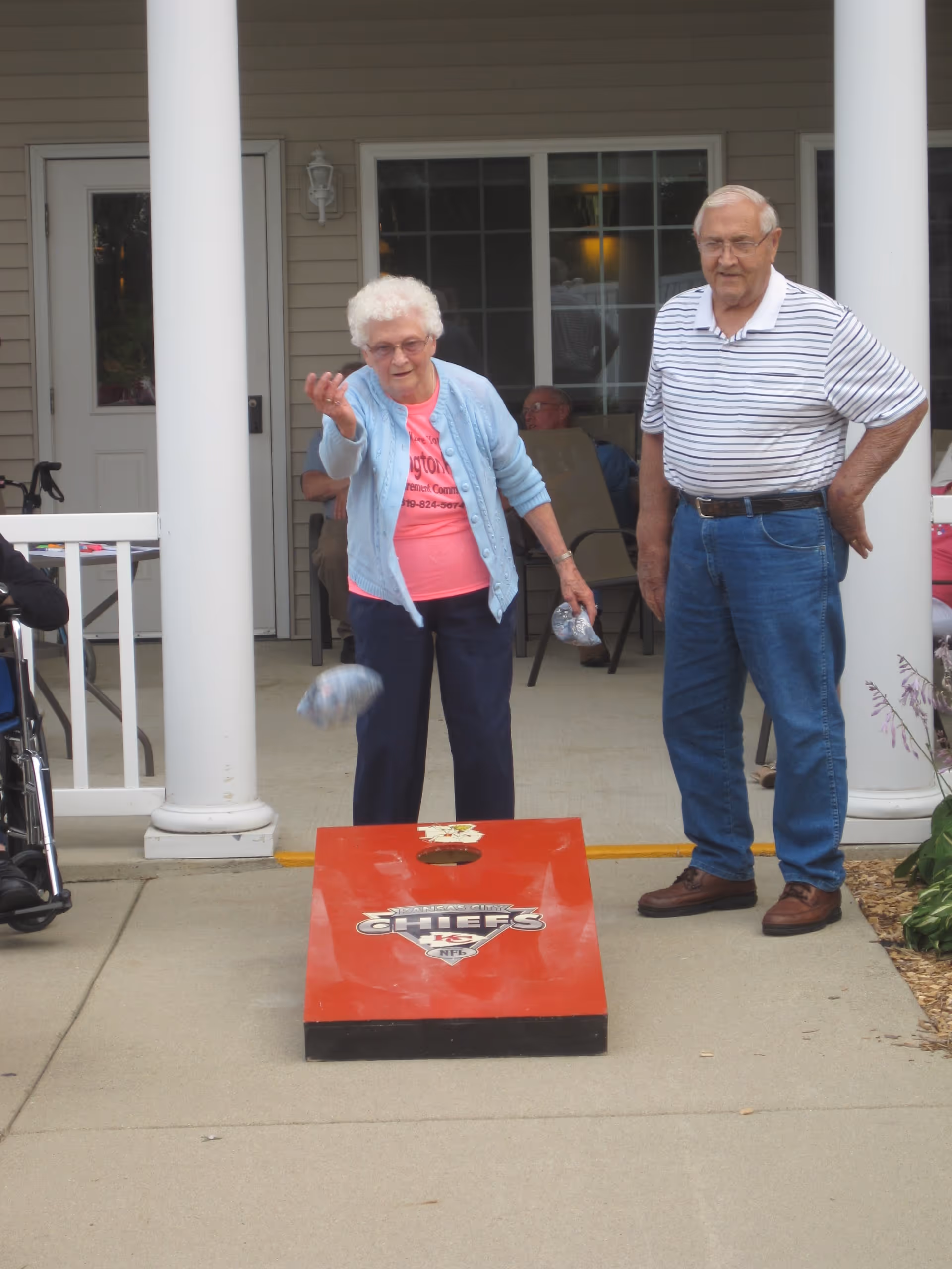An elderly woman and man are playing cornhole outside on a patio area. The woman is in the act of tossing a bean bag toward a red cornhole board with a Kansas City Chiefs logo. The man stands nearby watching. There are white pillars and a building entrance in the background.