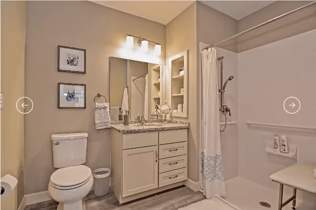 A clean and modern bathroom featuring a white toilet, a vanity with granite countertop and sink, a large mirror with three light fixtures above, built-in shelves with towels and toiletries, and a walk-in shower with a white curtain and a shower bench.