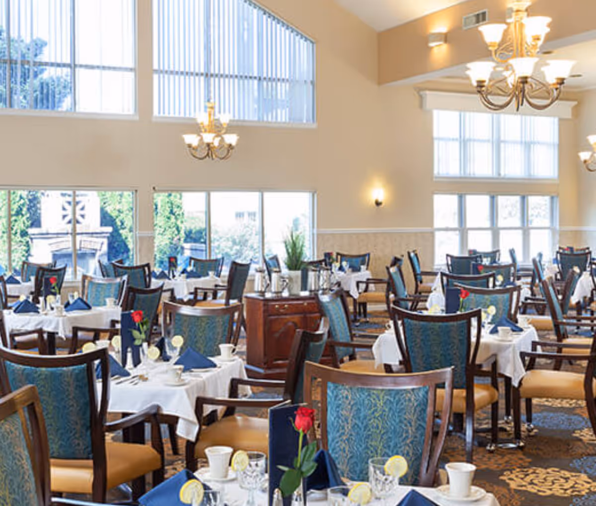 A bright and elegant dining room with multiple tables set with white tablecloths, blue napkins, cups, glasses with lemon slices, and single red roses in vases. The room features large windows with vertical blinds, high ceilings, chandeliers, and comfortable wooden chairs with blue patterned upholstery.